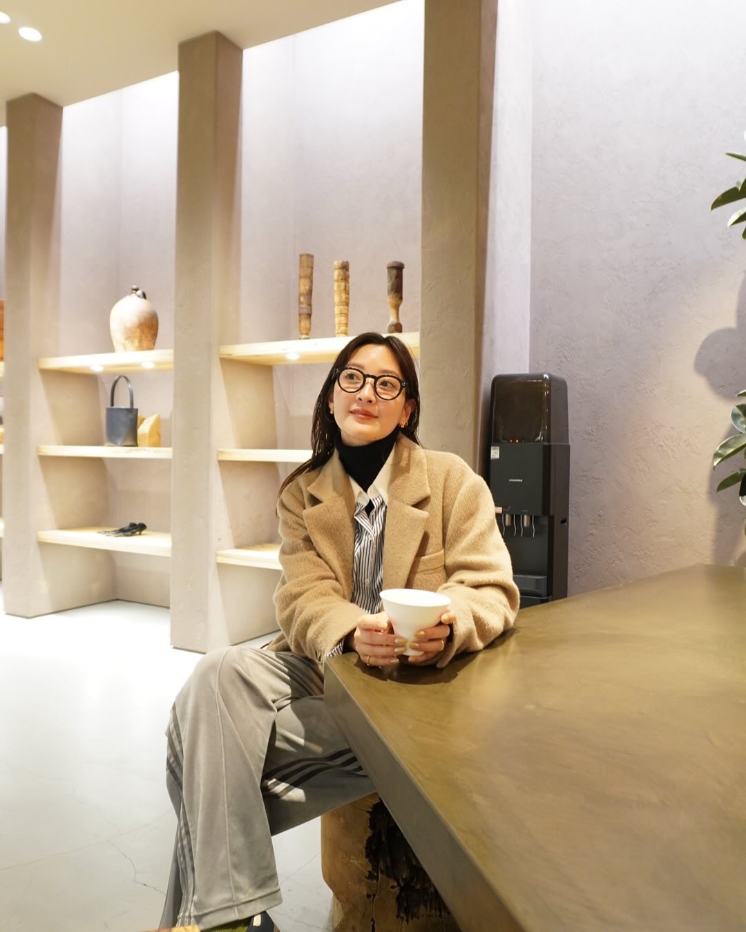 ベージュのコートを羽織り、メガネをかけてカフェでリラックスする知的なシャツスタイルの女性